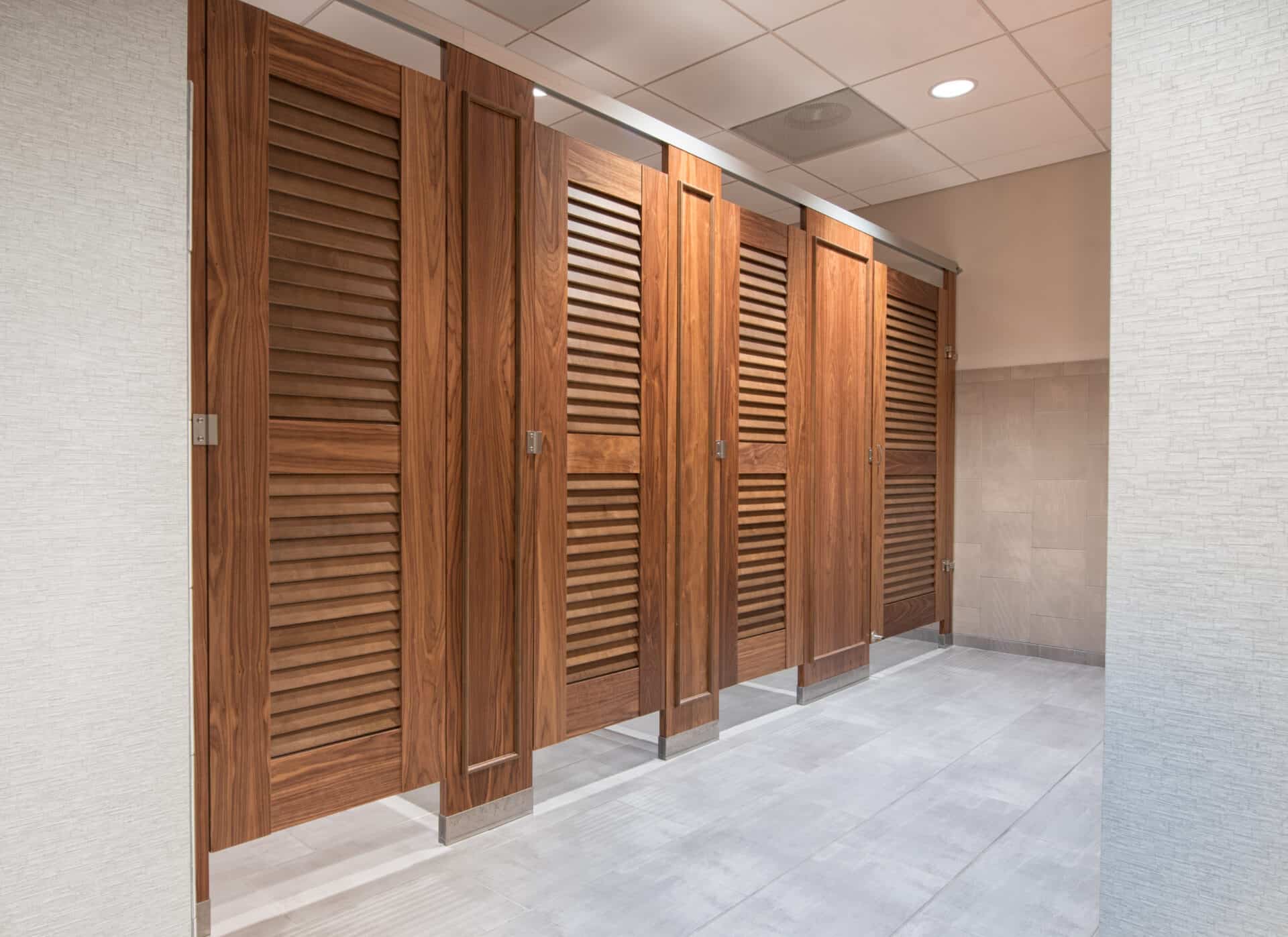 A row of four closed wood veneer restroom stalls with louvered doors headrail braced and moldings on pilasters in a public restroom facility.