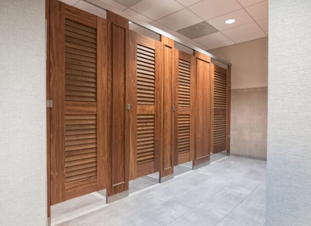 A row of four closed wood veneer restroom stalls with louvered doors headrail braced and moldings on pilasters in a public restroom facility.