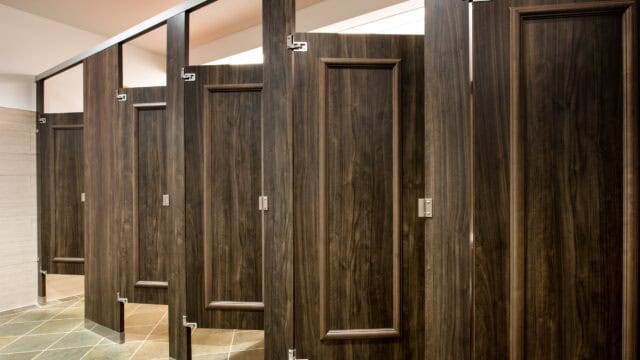 A commercial restroom with a row of partitions in dark wood veneer, headrail braced, with applied moldings on the doors.