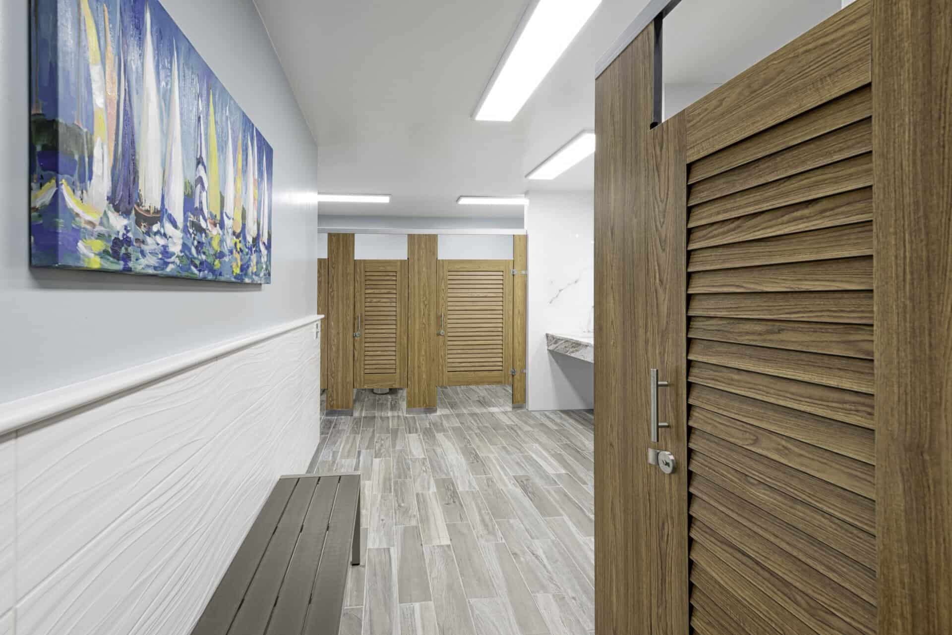Marina bathroom features compact laminate, wood grain louver doors in headrail braced style. Sailboats print on wall above grey metal changing bench.